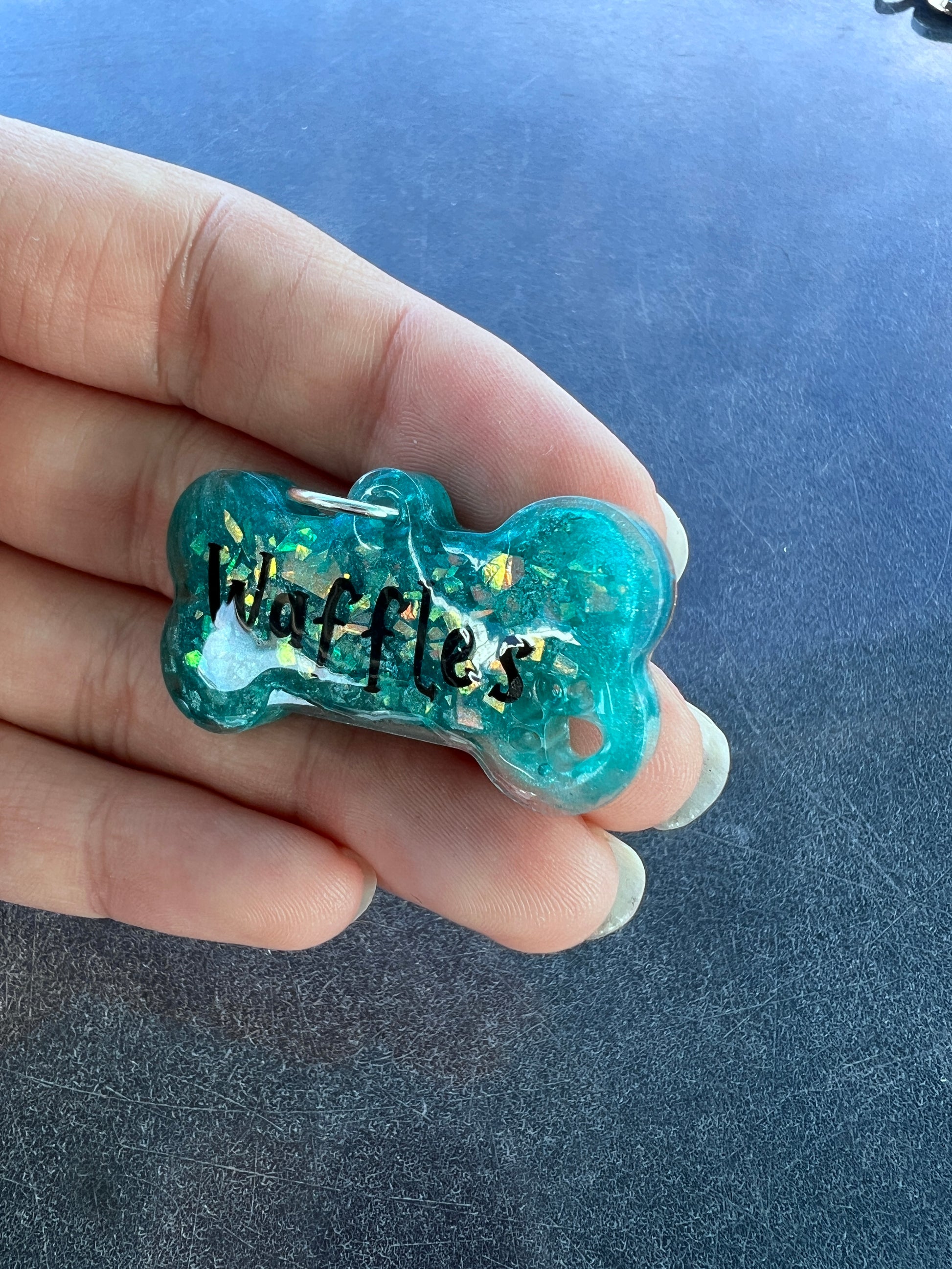 A vibrant teal bone-shaped resin pet play tag with the name 'Waffles' written in bold black text across the center.