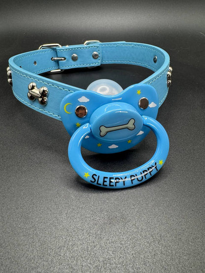 Blue dog collar straps with pacifier attachment that says Sleepy puppy with dog bone center and clouds and stars on shield on a gray background