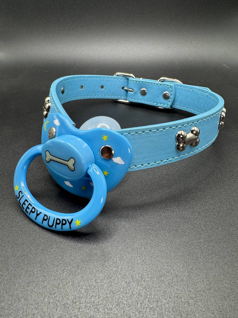 Blue dog collar straps with pacifier attachment that says Sleepy puppy with dog bone center and clouds and stars on shield on a gray background