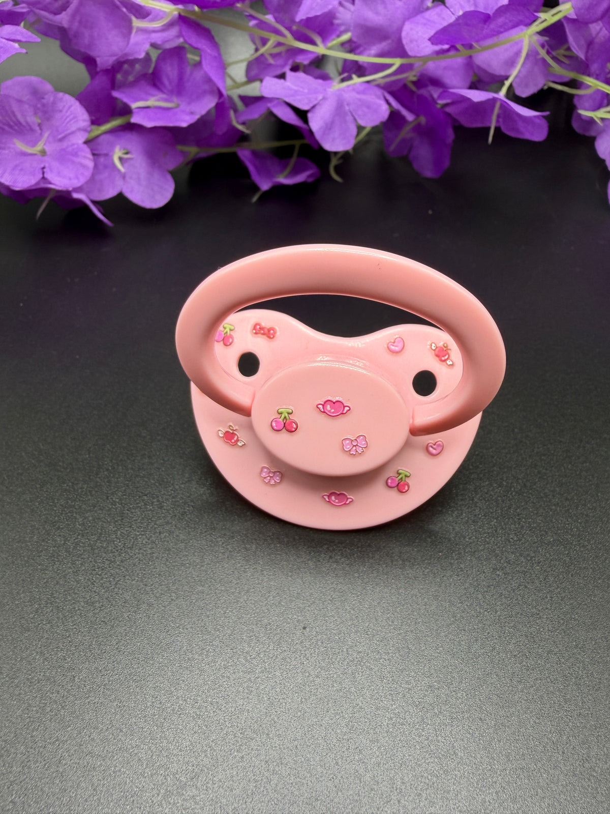 A pink age regress pacifier decorated with small cherry and bow motifs, displayed on a dark gray surface with purple flowers in the background.