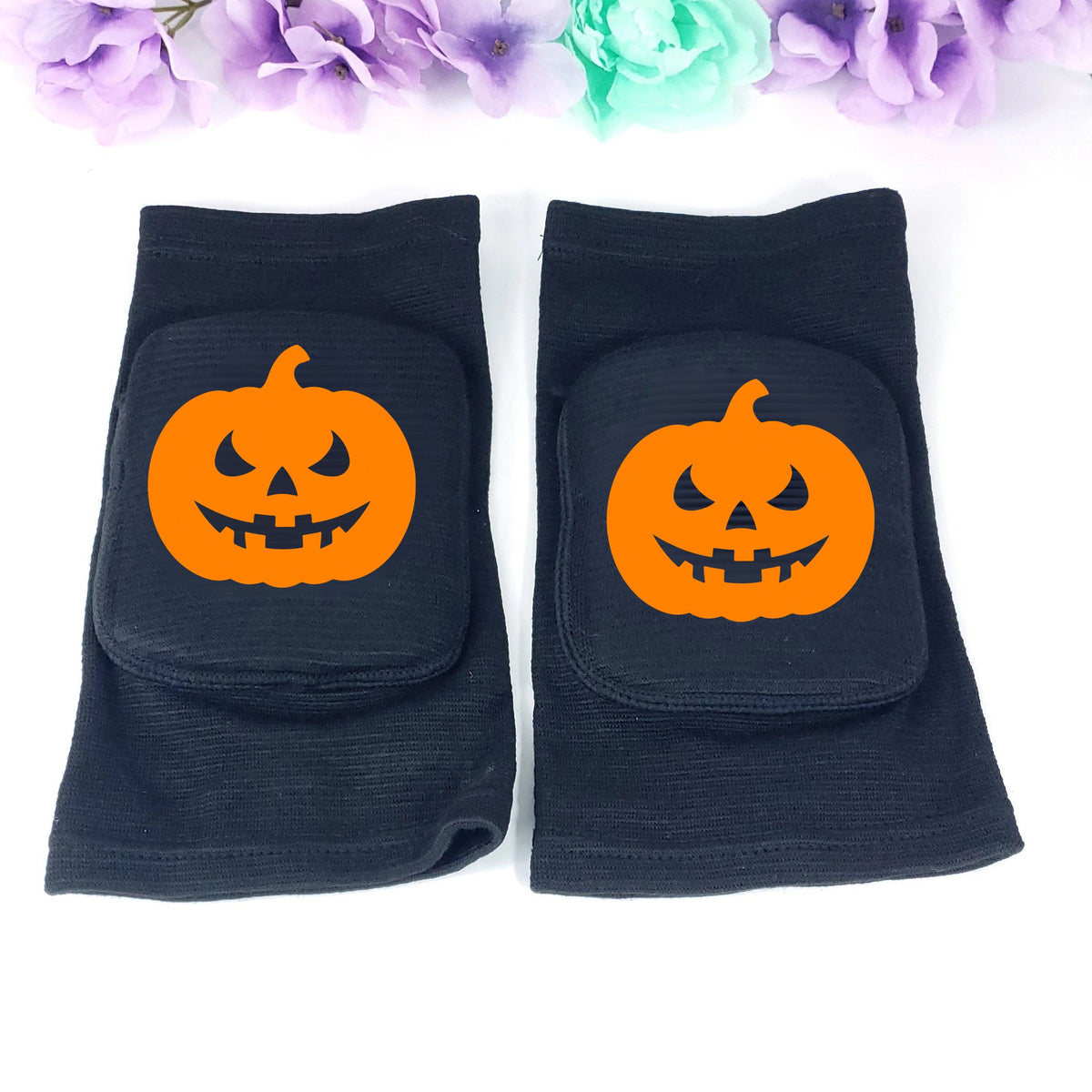 A pair of black knee pads with orange jack-o'-lantern pumpkin graphics on the front, displayed on a white background with decorative purple and mint flowers at the top.