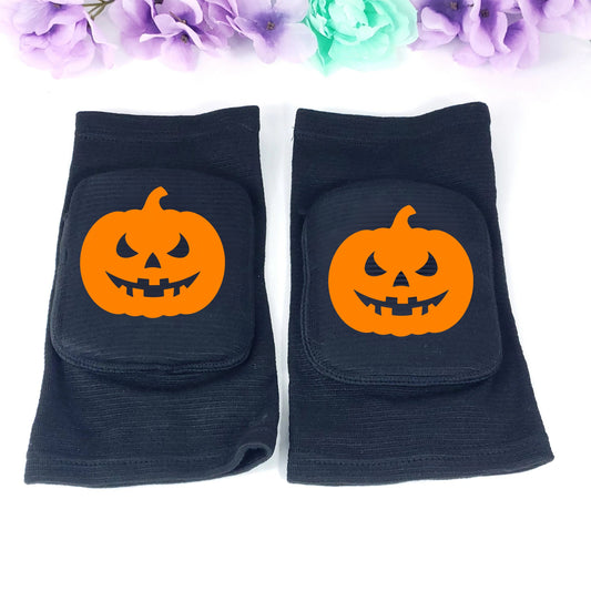 A pair of black knee pads with orange jack-o'-lantern pumpkin graphics on the front, displayed on a white background with decorative purple and mint flowers at the top.