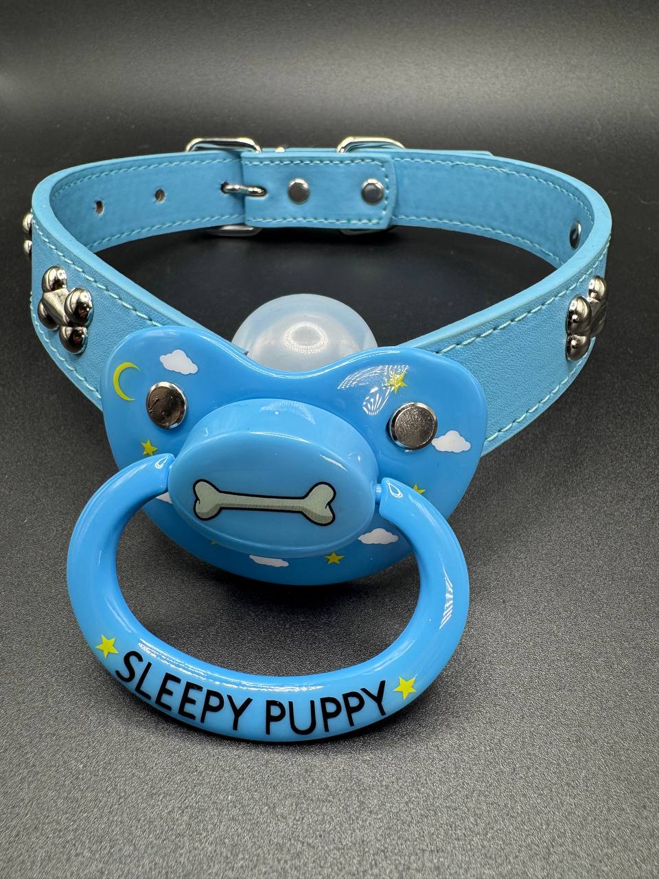 Blue dog bone straps with pacifier attachment with text 'Sleepy Puppy' on a gray background