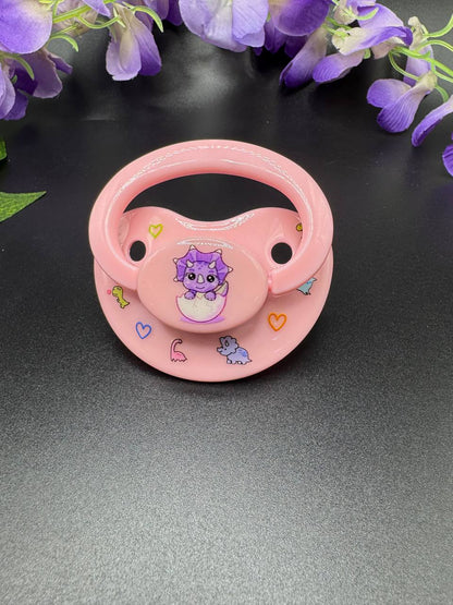 Purple triceratops image on pink ABDL adult pacifier with glow in the dark cute cartoon dinosaurs on the shield