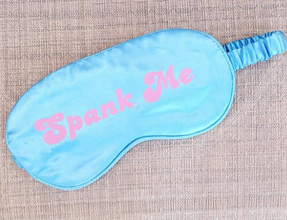 A blue silk adult blindfold with the text 'Spank Me' printed in pink on the front, featuring an elastic strap.