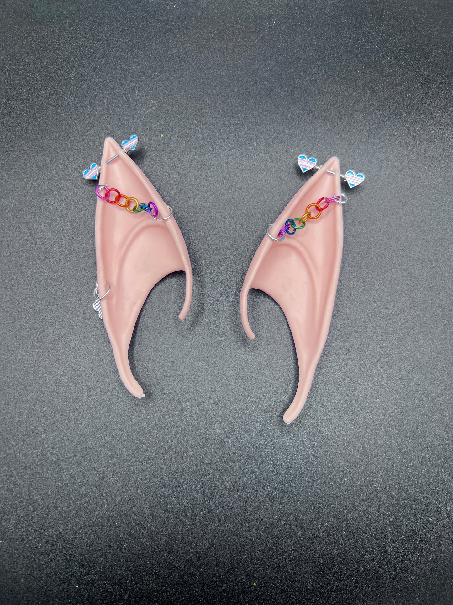 Pair of tan elf ears with trans pride hearts bar earrings and rainbow chain going across earsd a gray background