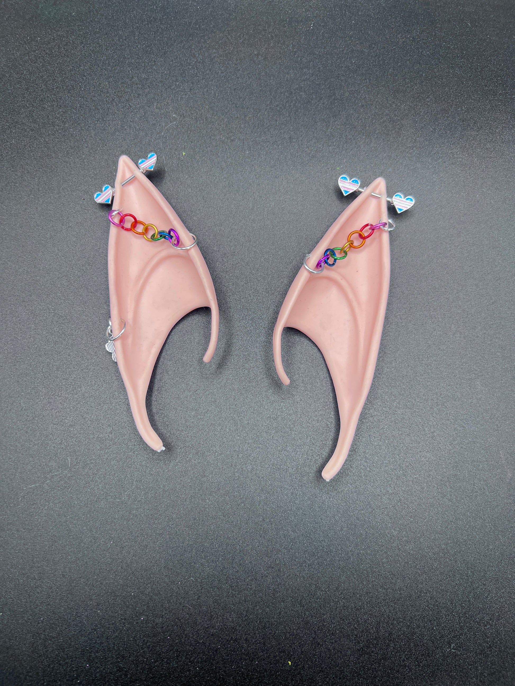 Pair of tan elf ears with trans pride hearts bar earrings and rainbow chain going across earsd a gray background