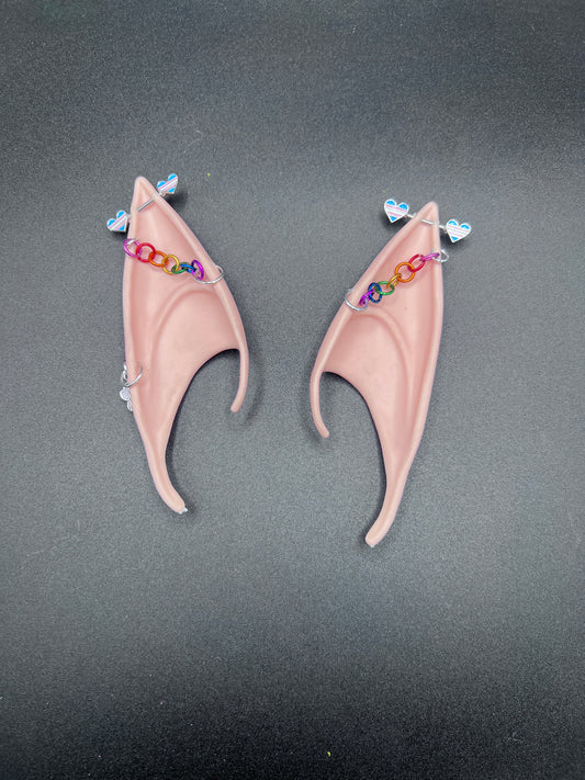 Pair of tan elf ears with trans pride hearts bar earrings and rainbow chain going across earsd a gray background