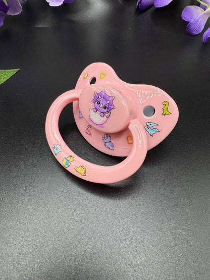 baby triceratops coming out of shell image on button of adult pacifier with glow in the dark dinosaurs and hearts on shield and handle