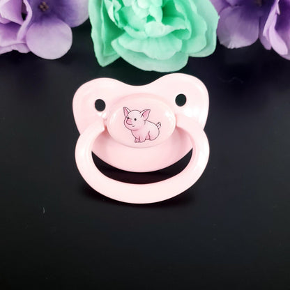 Pink adult pacifier with a pig illustration on a black background with flowers.