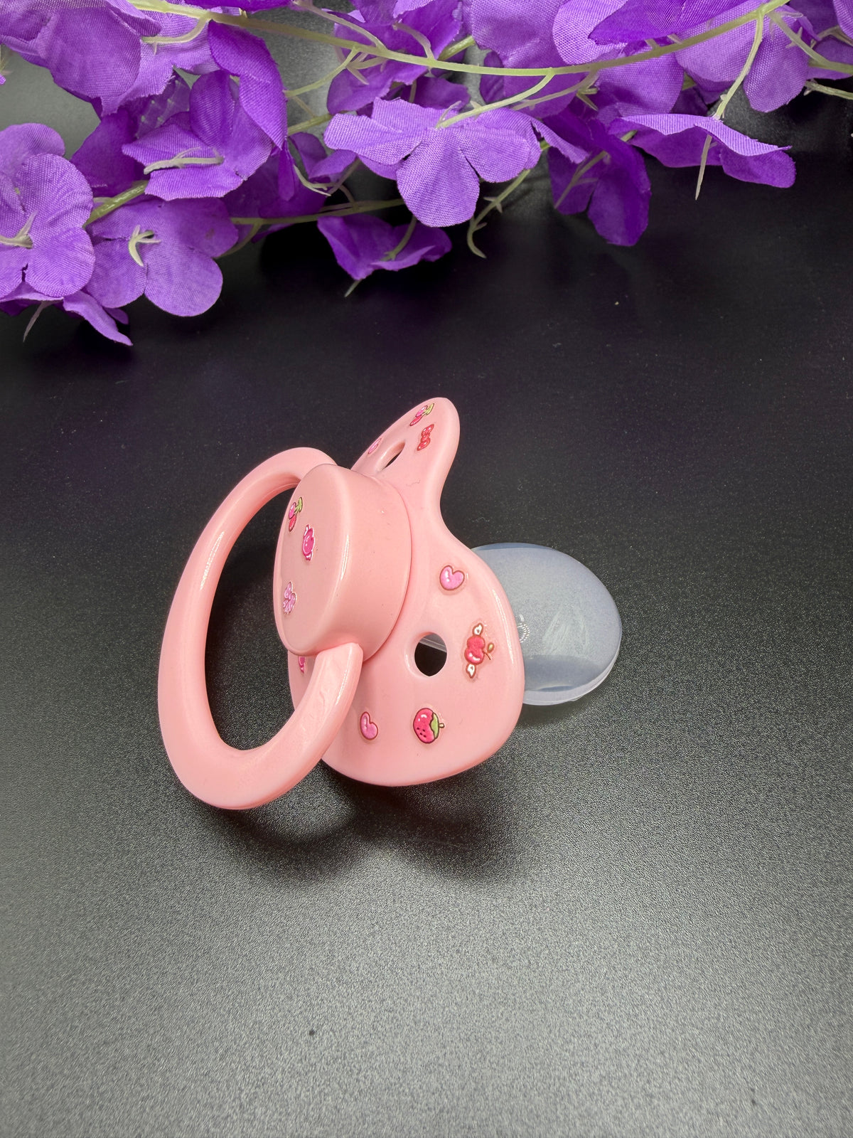 A pink baby pacifier with a matching ring and a clear silicone shield, decorated with small cherry and heart details, resting on a dark gray surface with purple flowers in the background.