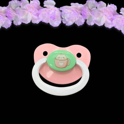 An adult pacifier with a cocoa cat design, featuring a white handle and  a marshmallow cat illustration and text on a mint button.