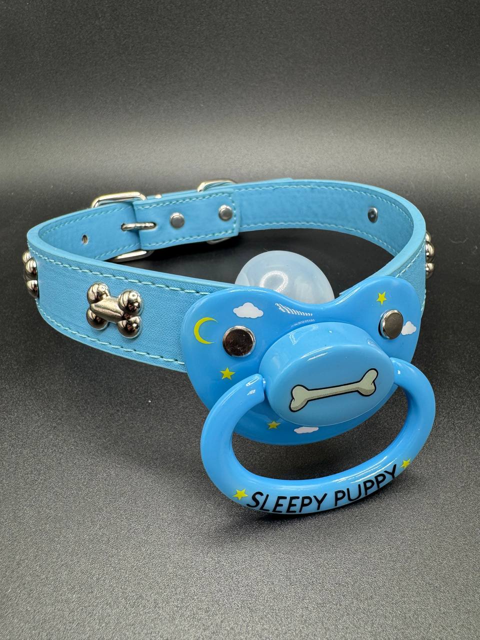 Blue gag straps with pacifier attachment that says Sleepy puppy with dog bone center and clouds and stars on shield on a gray background