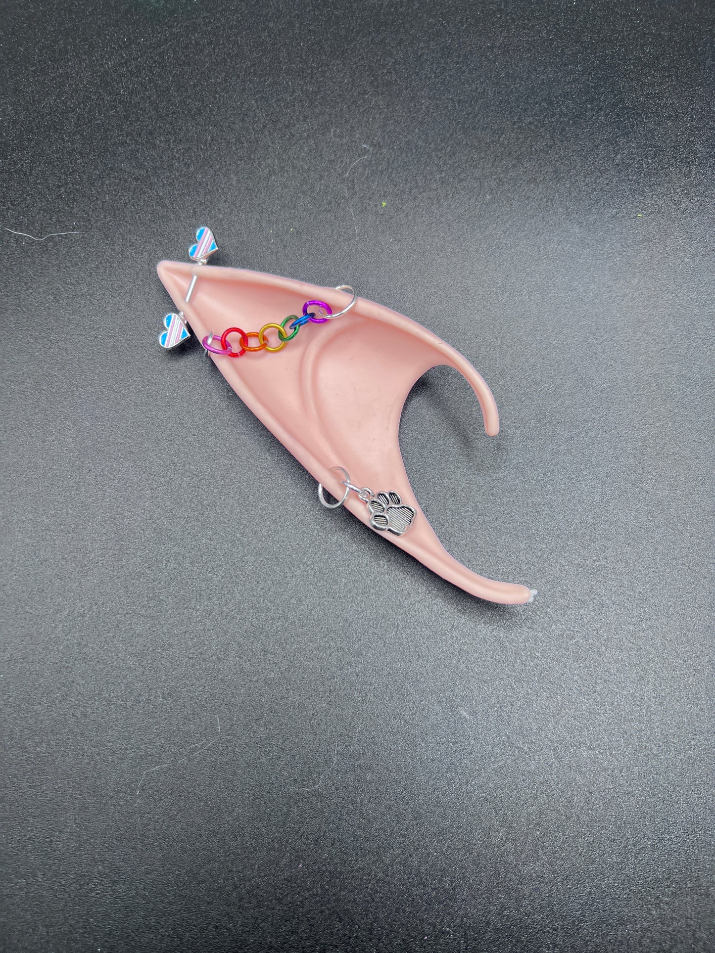 fairy elf ear with paw charm earring, rainbow chain going across and barbell trans hearts piercing at the tip