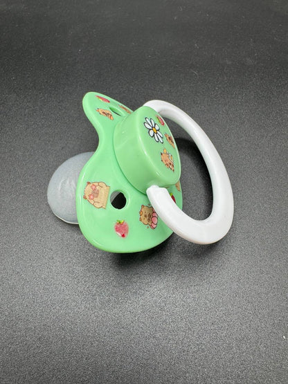 Green adult binkie pacifier with floral and capybara designs on a gray background