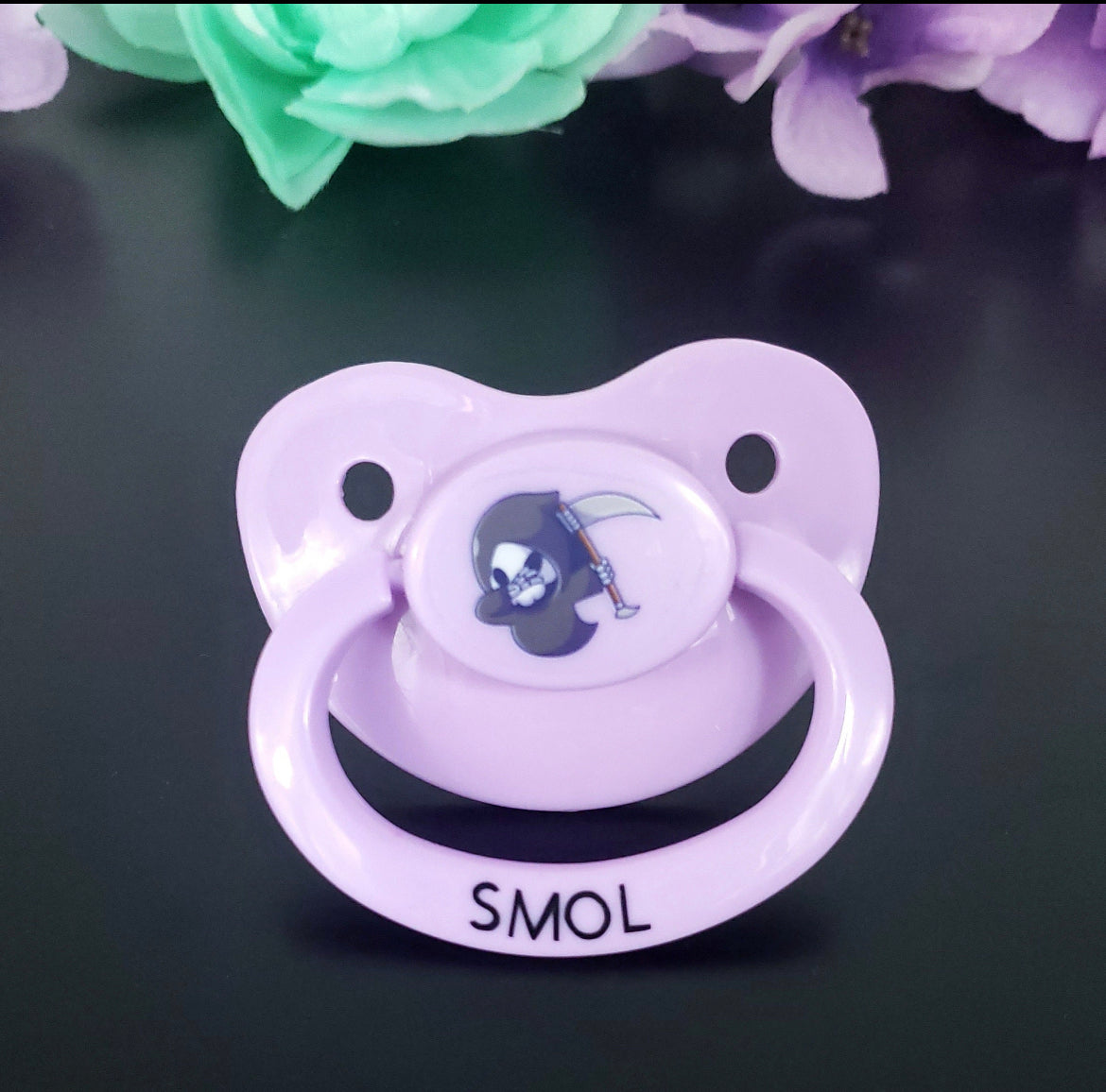 lavender adult pacifier with kawaii style grim reaper dabbing center. Text on the handle saying Smol in black lettering