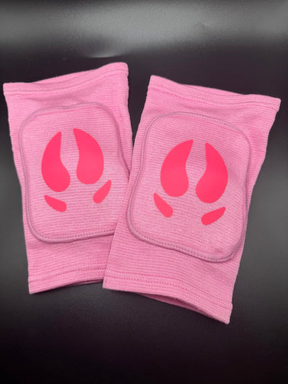 A pair of pink ribbed knee protectors with a bright pink stylized pig hooves on the front, displayed on a dark surface.