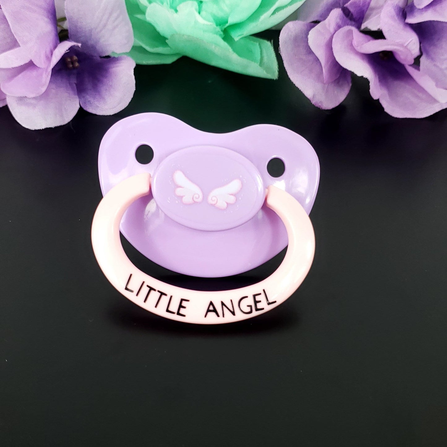 lavender adult pacifier with baby pink handle. Center button features cute pink angel wings and the handle says little angel