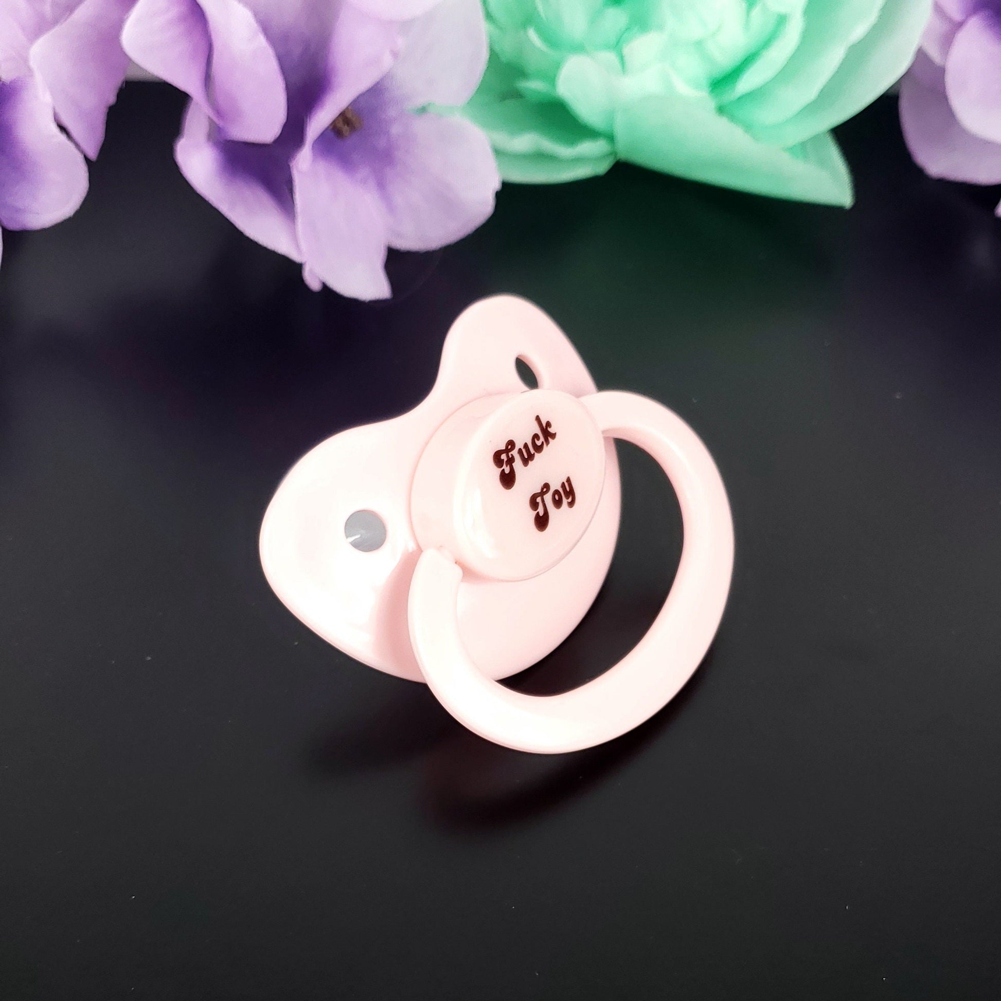 A baby pink adult pacifier with bold text in the center that reads "Fuck Toy," featuring a mature and edgy design.