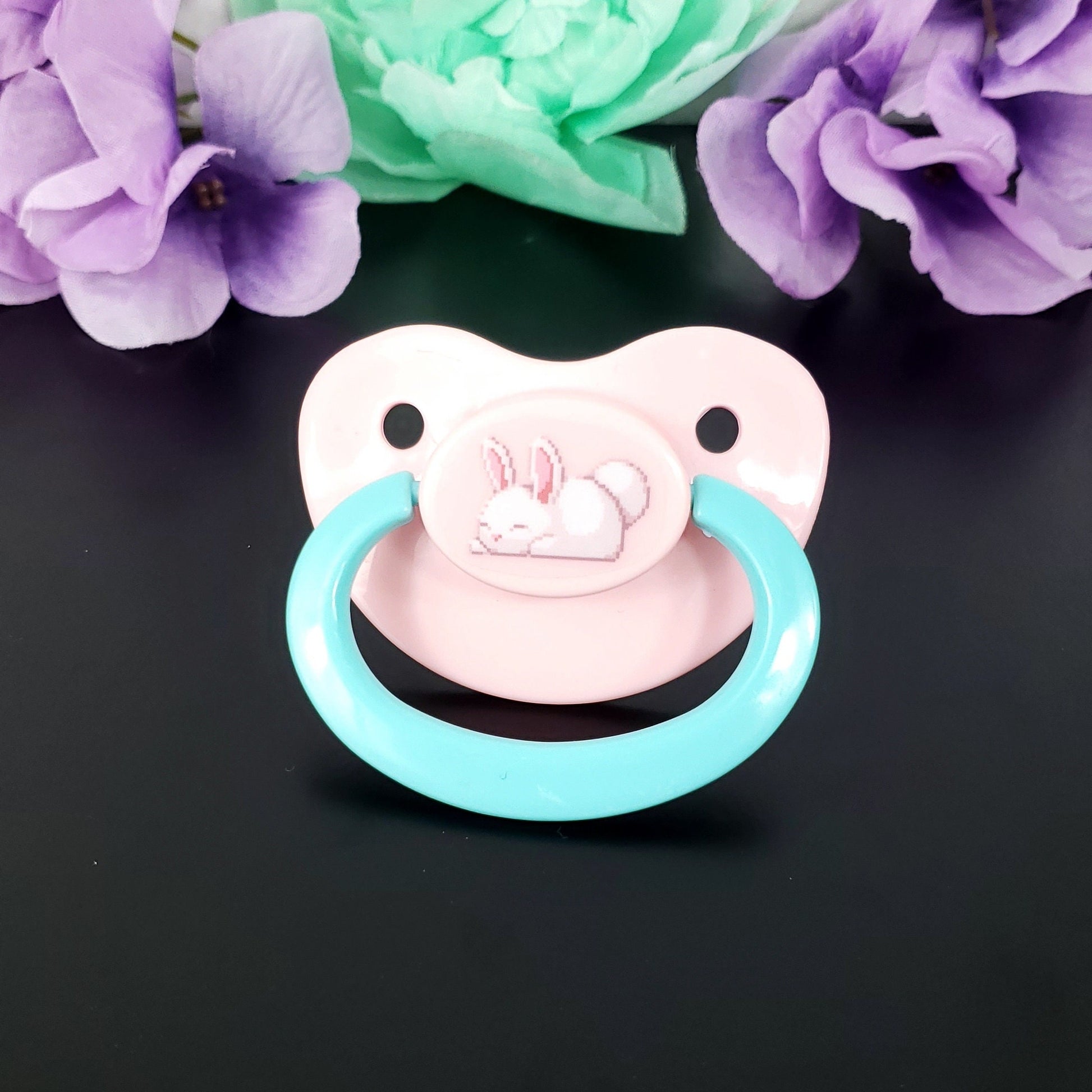 Pastel pink adult pacifier ith aqua handle featuring a cute sleeping white bunny