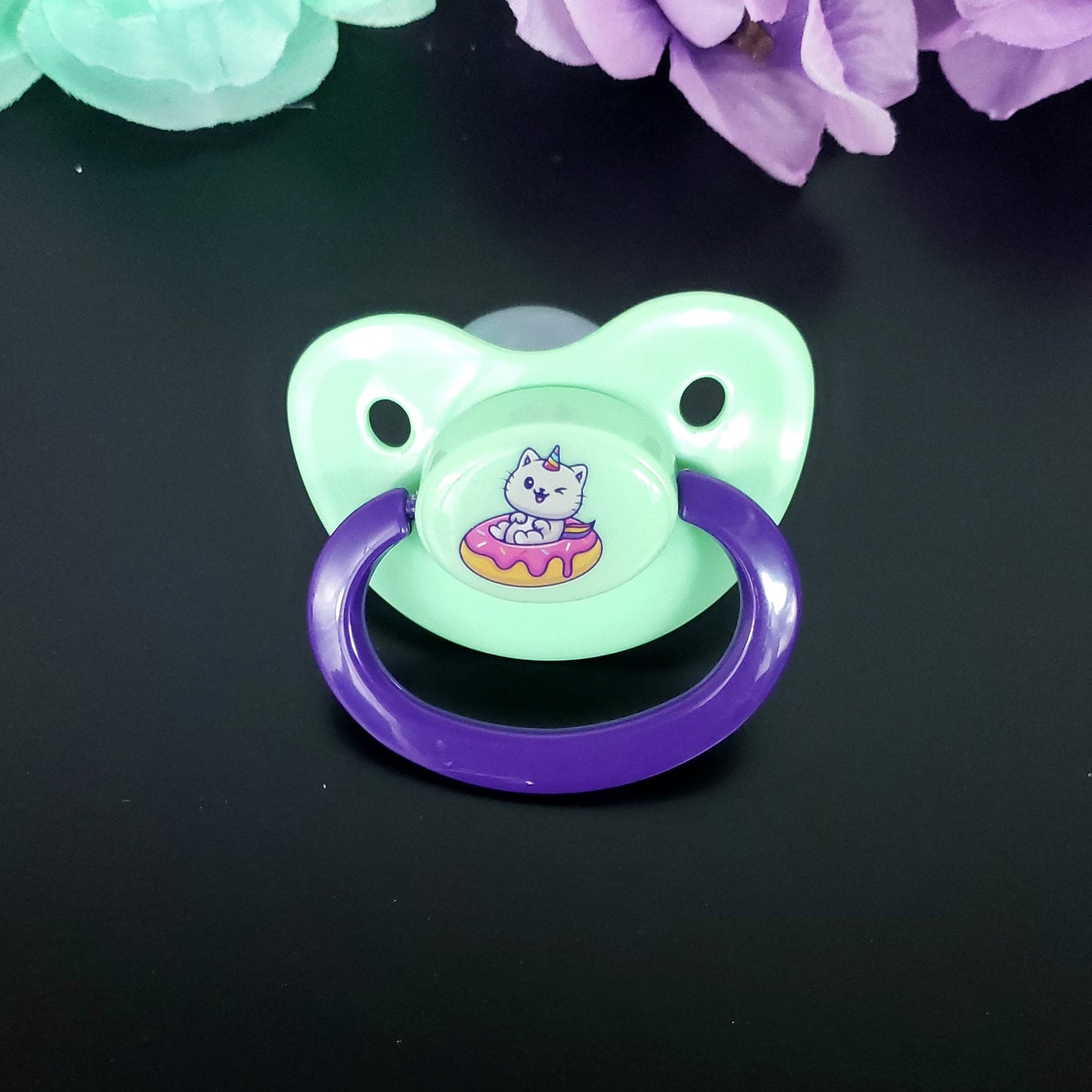 Mint adult pacifier with dark purple handle featuring a fun kawaii white cat unicorn with rainbow horn and tail, sitting in a frosted donut with sprinkles