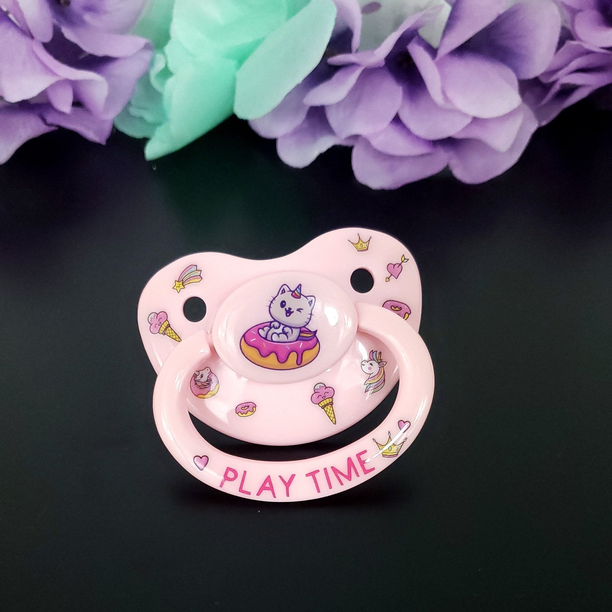Baby pink Adult deco pacifier with a unicorn cat sitting in a frosted sprinkle donut in the center. The shield is decorated with images of unicorns, ice cream, crowns and donuts. The handle says play time in hot pink with a crown and hearts on each side.
