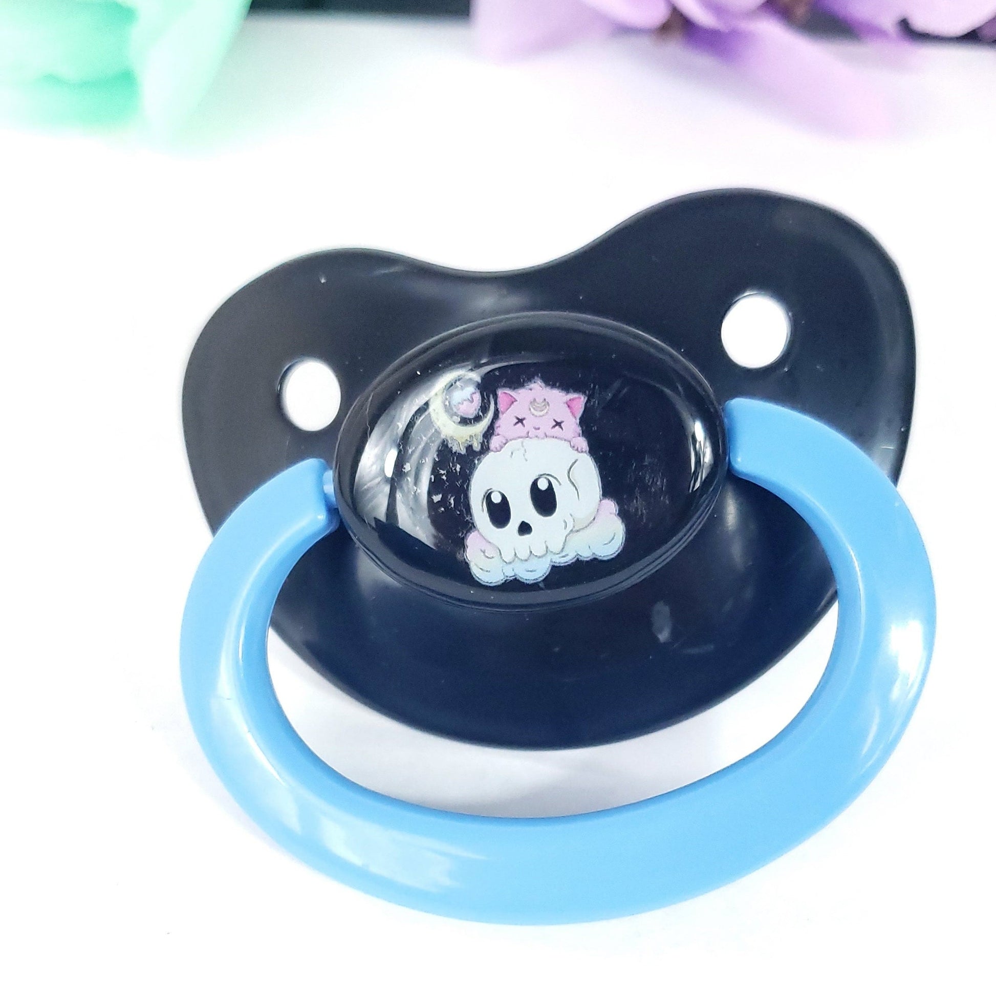 black adult pacifier with baby blue handle featuring a creepy kawaii cat peaking over a skull with dripping moon and pastel clouds