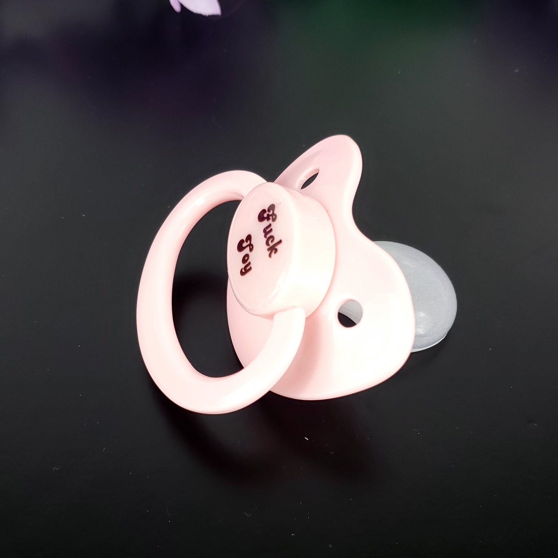 A baby pink adult pacifier with bold text in the center that reads "Fuck Toy," featuring a mature and edgy design.