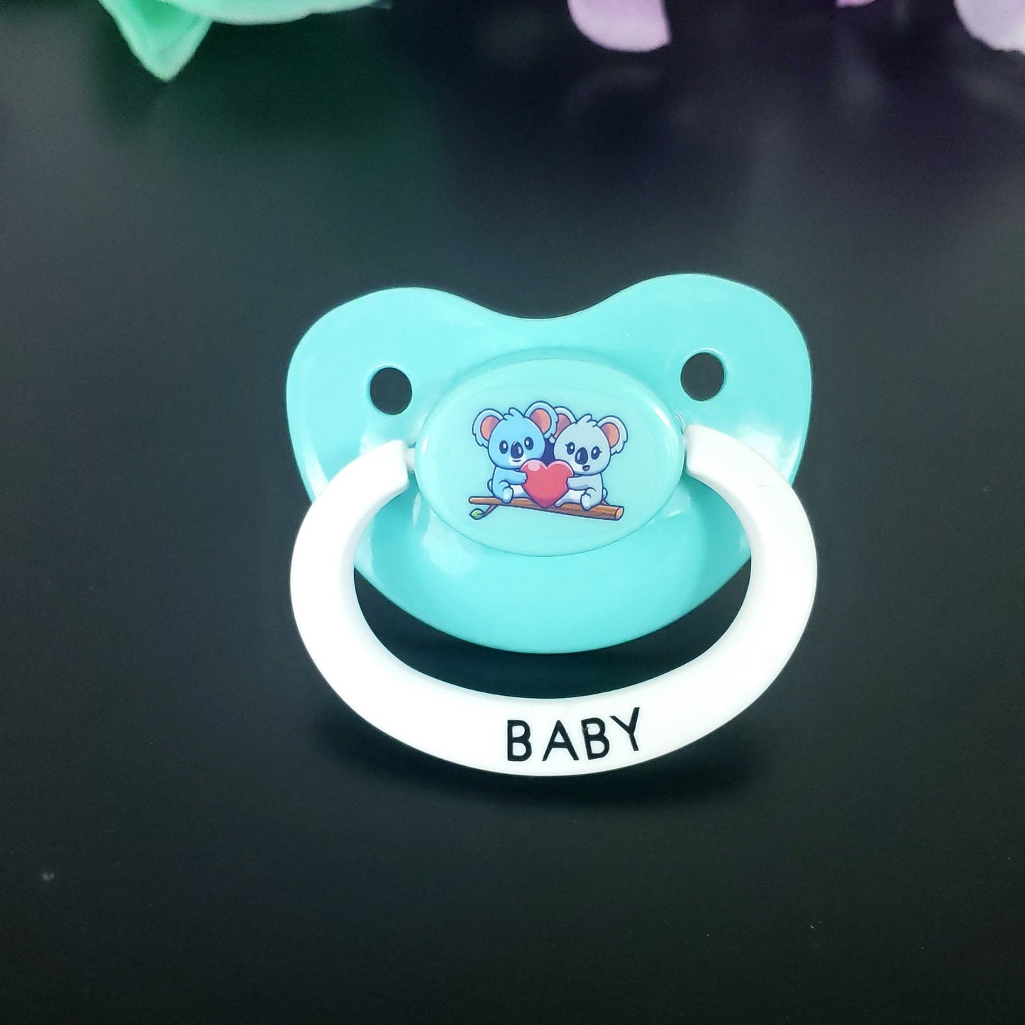 aqua adult pacifier with two koalas hugging a heart. Pacifier handle is white and has text that says baby