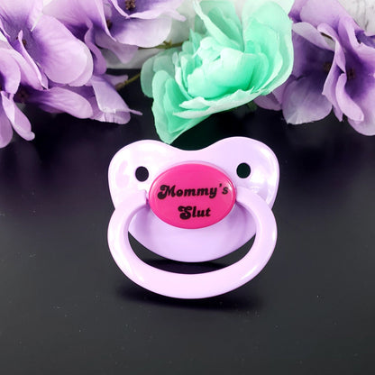 A lavender and hot pink adult pacifier with bold text in the center that reads "Mommy's Slut," featuring a vibrant and edgy design.