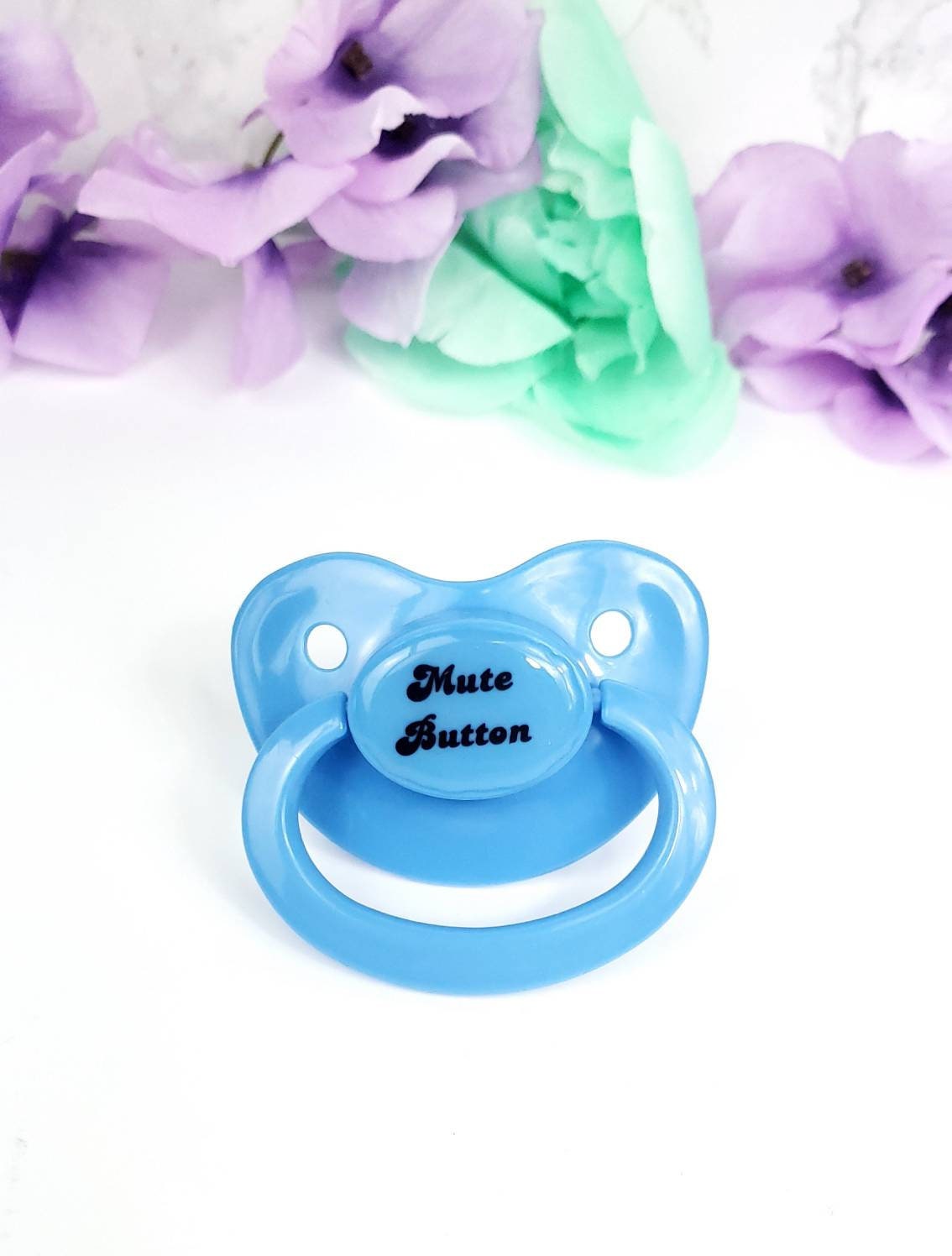 A baby blue adult pacifier with elegant black text in the center that reads "Mute Button," featuring a soft and playful design.