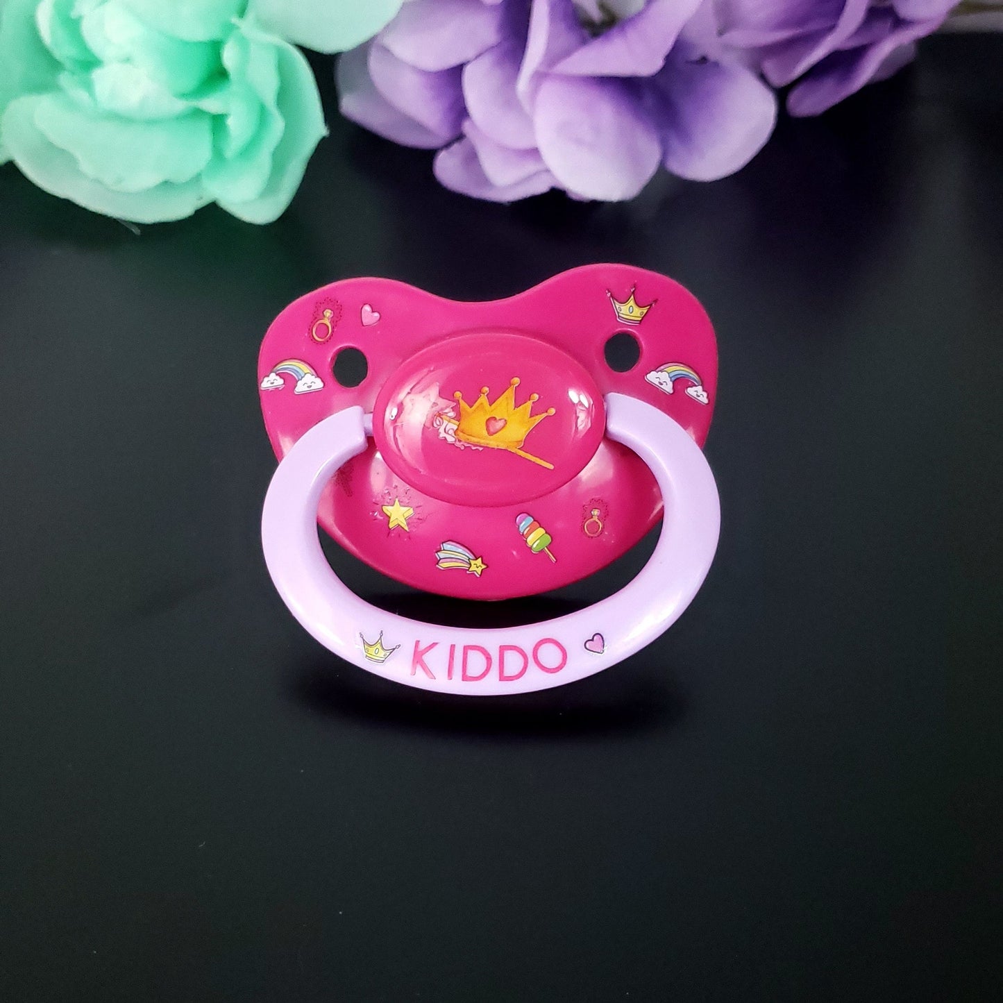 Hot pink adult pacifier with lavender handle featuring a crown and want in the center. On the shield is playful princess items such as diamond rings, crowns, stars, rainbows and hearts. The handle has hot pink text saying kiddo with a heart and crown on each side of text