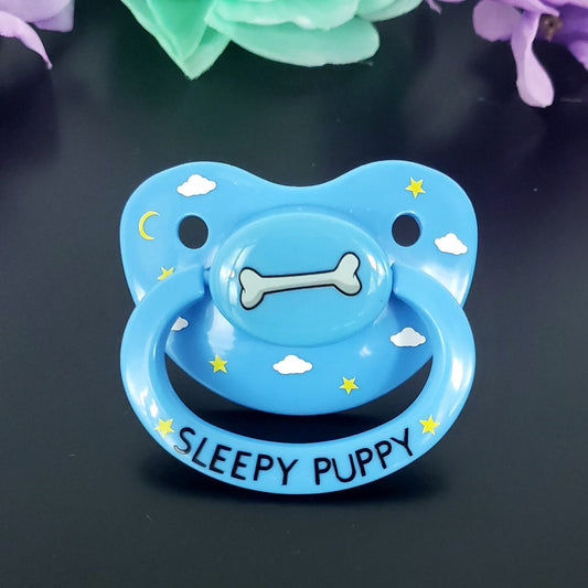 A blue adult pacifier with a sleepy puppy design, featuring stars and clouds and the text 'SLEEPY PUPPY' on the handle.