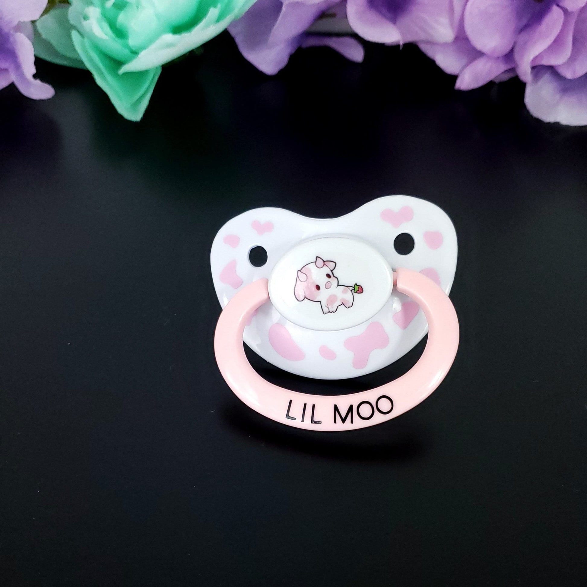 White adult pacifier with baby pink handle. Center button has kawaii strawberry pink cow and the shield has baby pink cow spots with hearts. Then the handle says Lill Moo