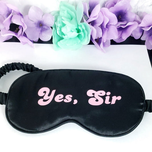 A black adult blindfold with the text 'YES, SIR' printed in pink on the front, displayed on a flat surface with decorative flowers around it.