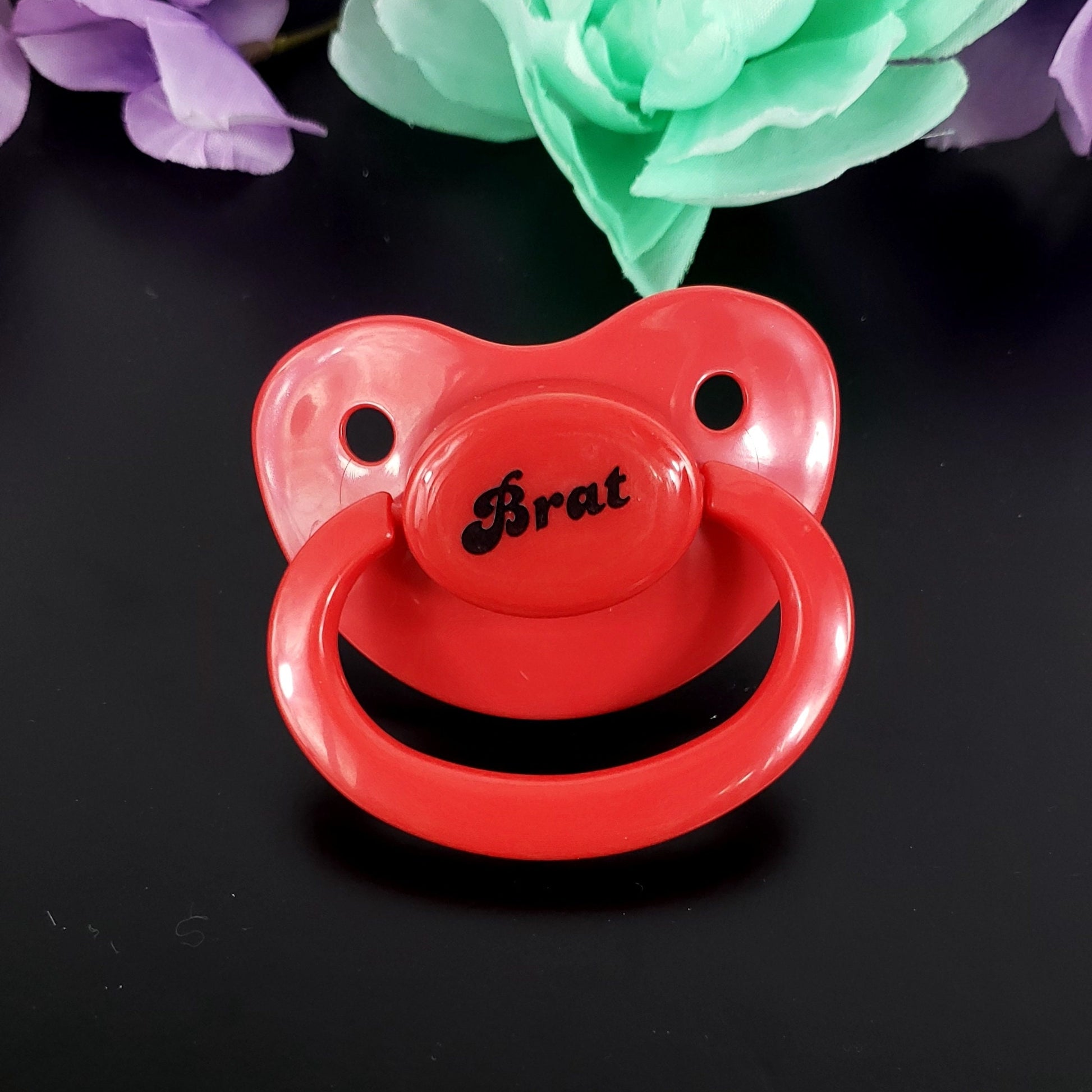 A red adult pacifier featuring the word "Brat" boldly displayed in the center, designed for playful and cheeky self-expression.