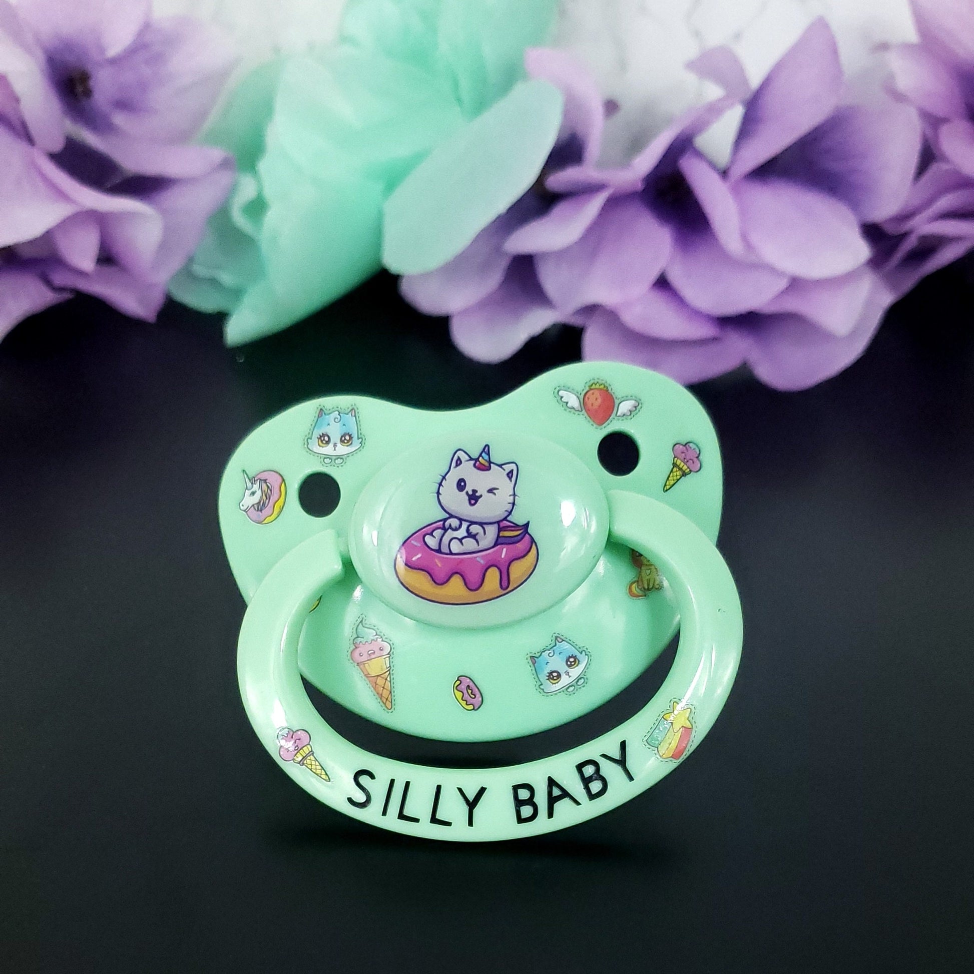 mint adult pacifier featuring a cat unicorn sitting in a frosted donut on the button. Surrounding on the shield is cute images of donuts, ice cream, blue kittens, flying strawberry and unicorns. On the handle it says silly baby with an image of ice cream and rainbow star on each side.