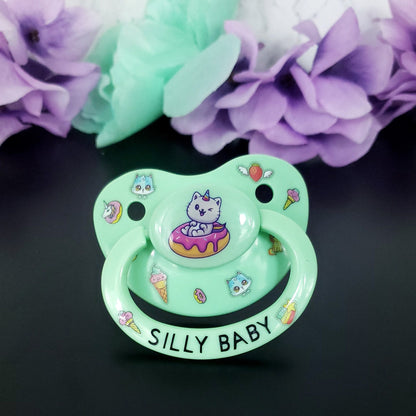 mint adult pacifier featuring a cat unicorn sitting in a frosted donut on the button. Surrounding on the shield is cute images of donuts, ice cream, blue kittens, flying strawberry and unicorns. On the handle it says silly baby with an image of ice cream and rainbow star on each side.