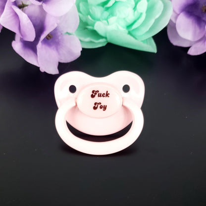 A baby pink adult pacifier with bold text in the center that reads "Fuck Toy," featuring a mature and edgy design.