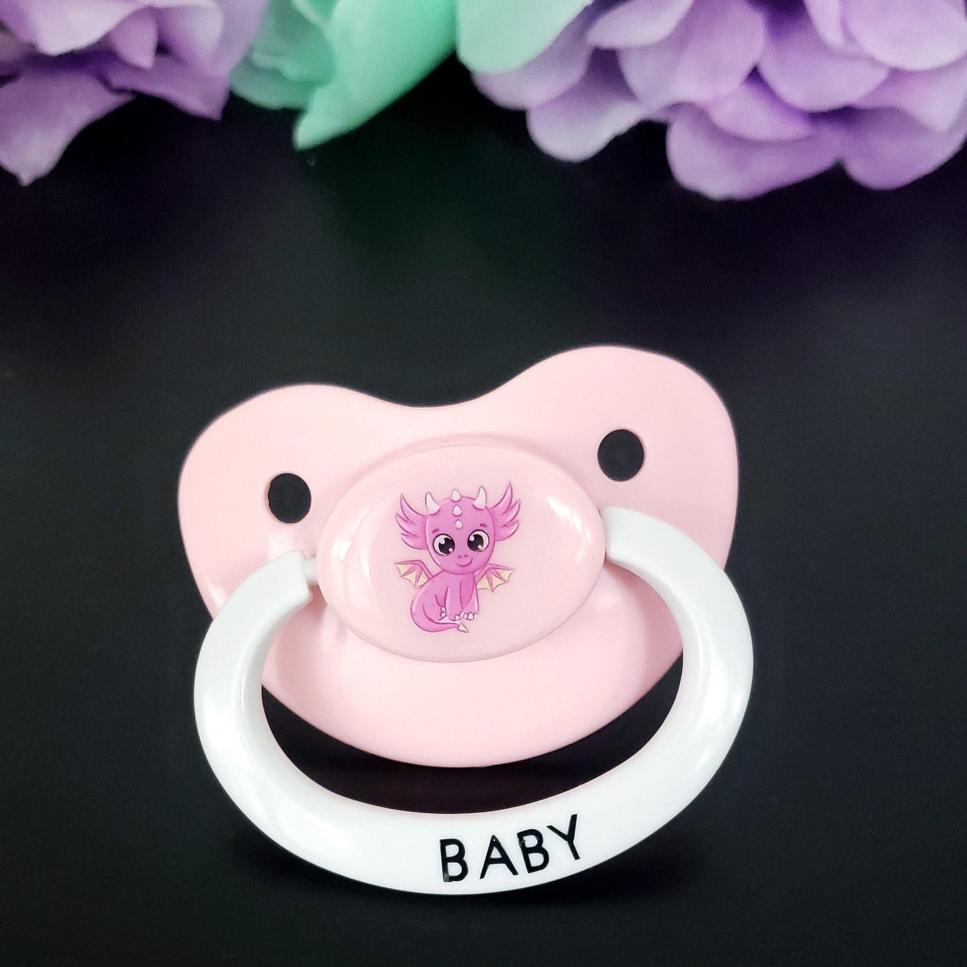 baby pink adult pacifier with white handle featuring a cute hot pink dragon in the center. The handle has black text that says baby.