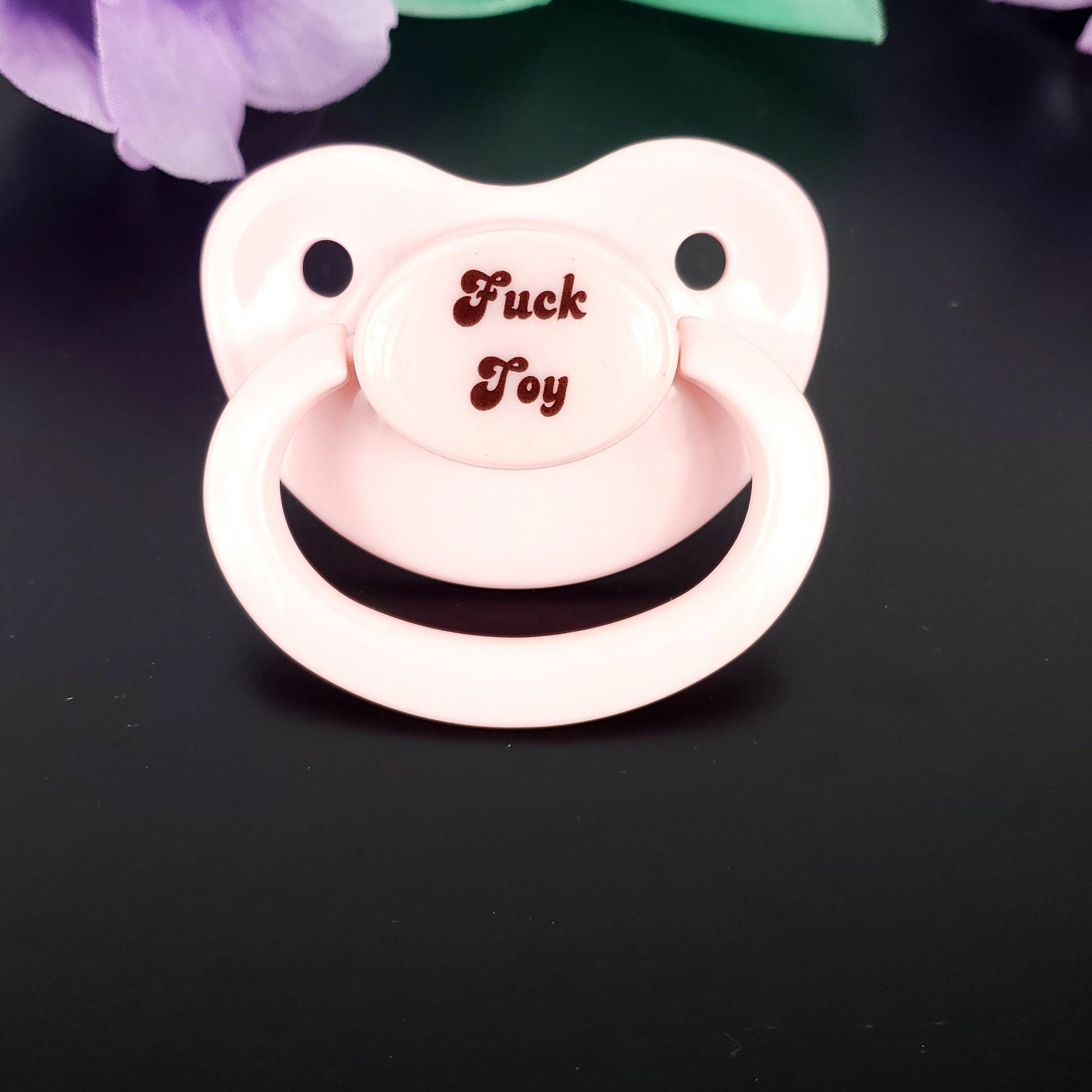 A baby pink adult pacifier with bold text in the center that reads "Fuck Toy," featuring a mature and edgy design.