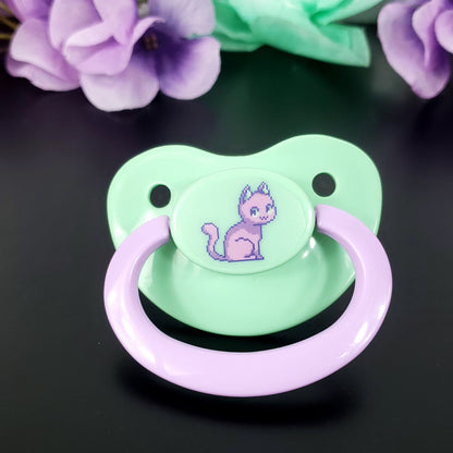 mint adult pacifier with lavender handle. Featuring a purple pixel cat in the center.